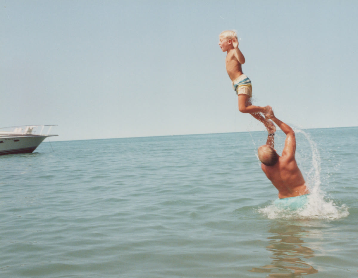 A boy launched high into the air off his father's hands, out in the lake on a summer day. The father's back is to the camera.
